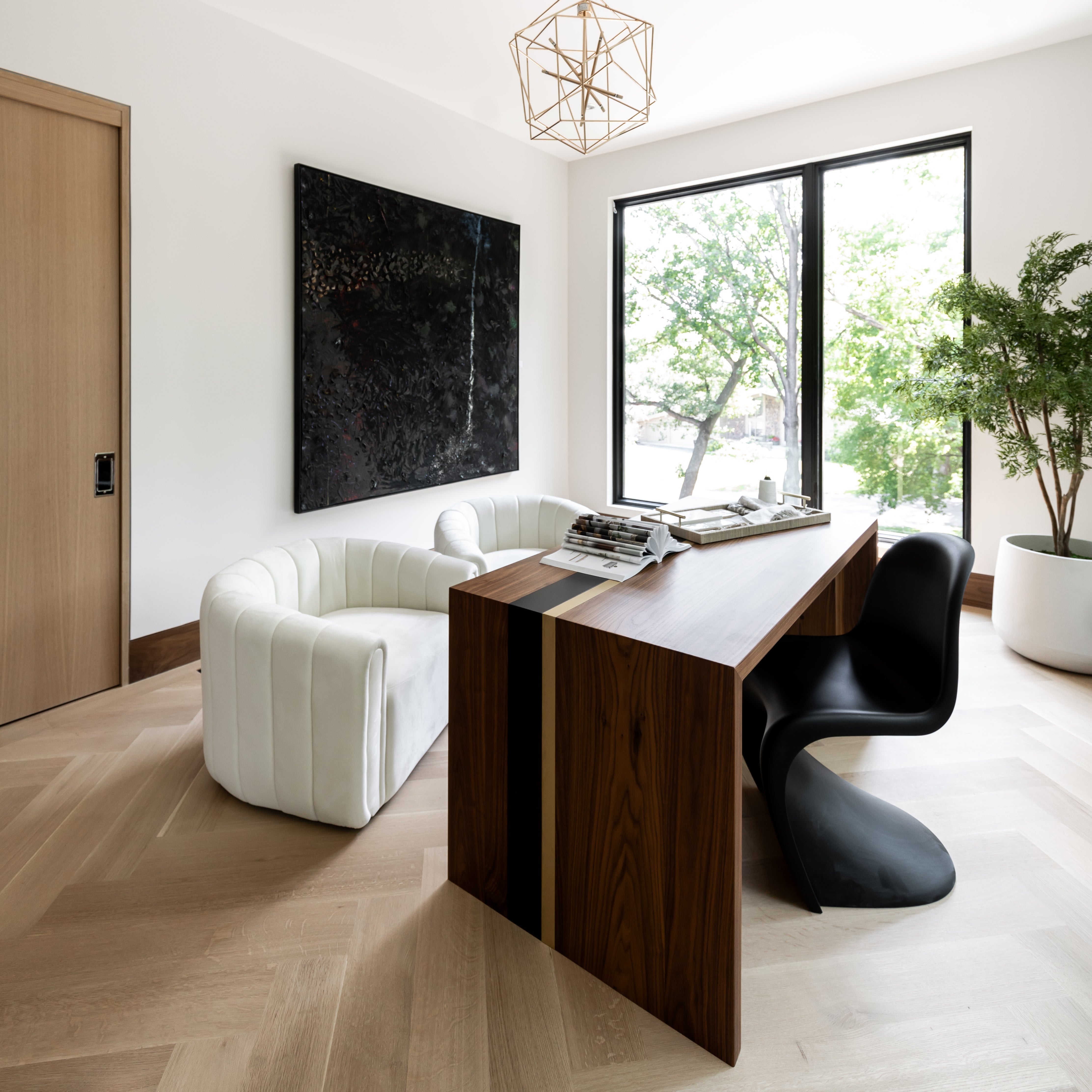 A modern home office desk with a dark wood finish, paired with a white upholstered chair and a black office chair. The room has a minimalist design with a large window, a decorative chandelier, and plants in the background.